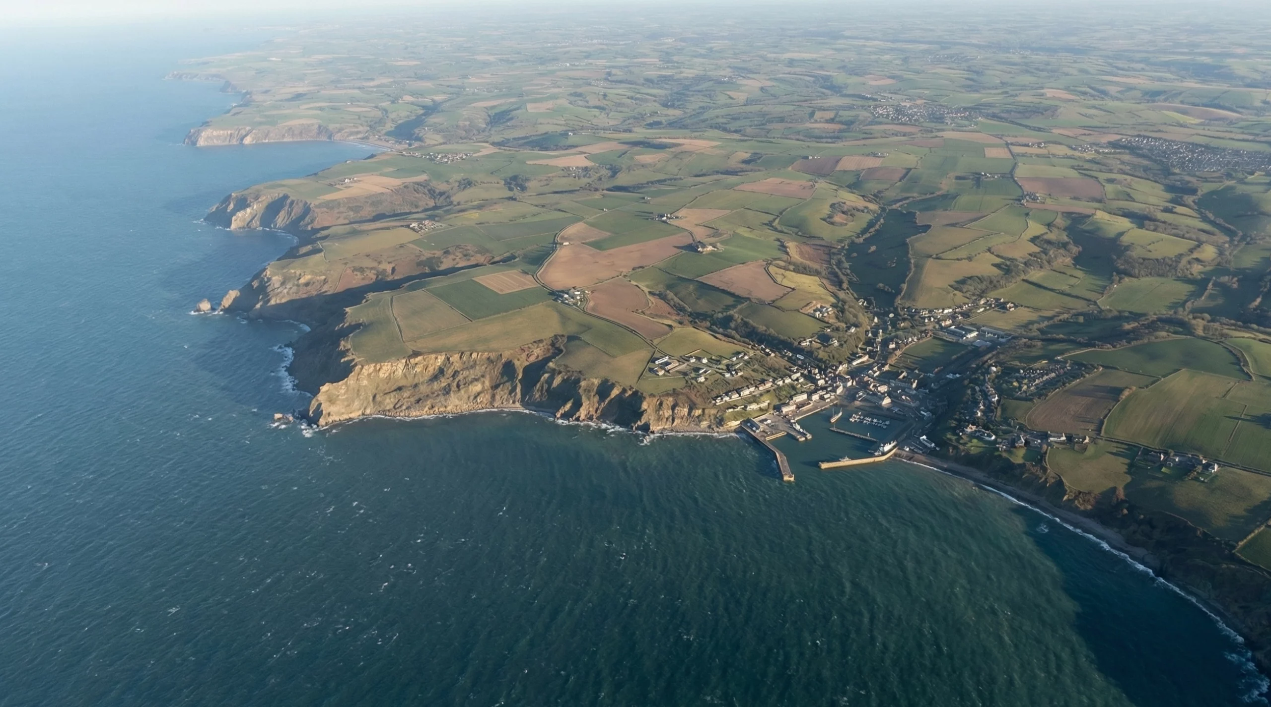 Vue aérienne oblique d'un littoral escarpé avec des terres agricoles et un port de plaisance, capturée à haute altitude pour l'observation terrestre.