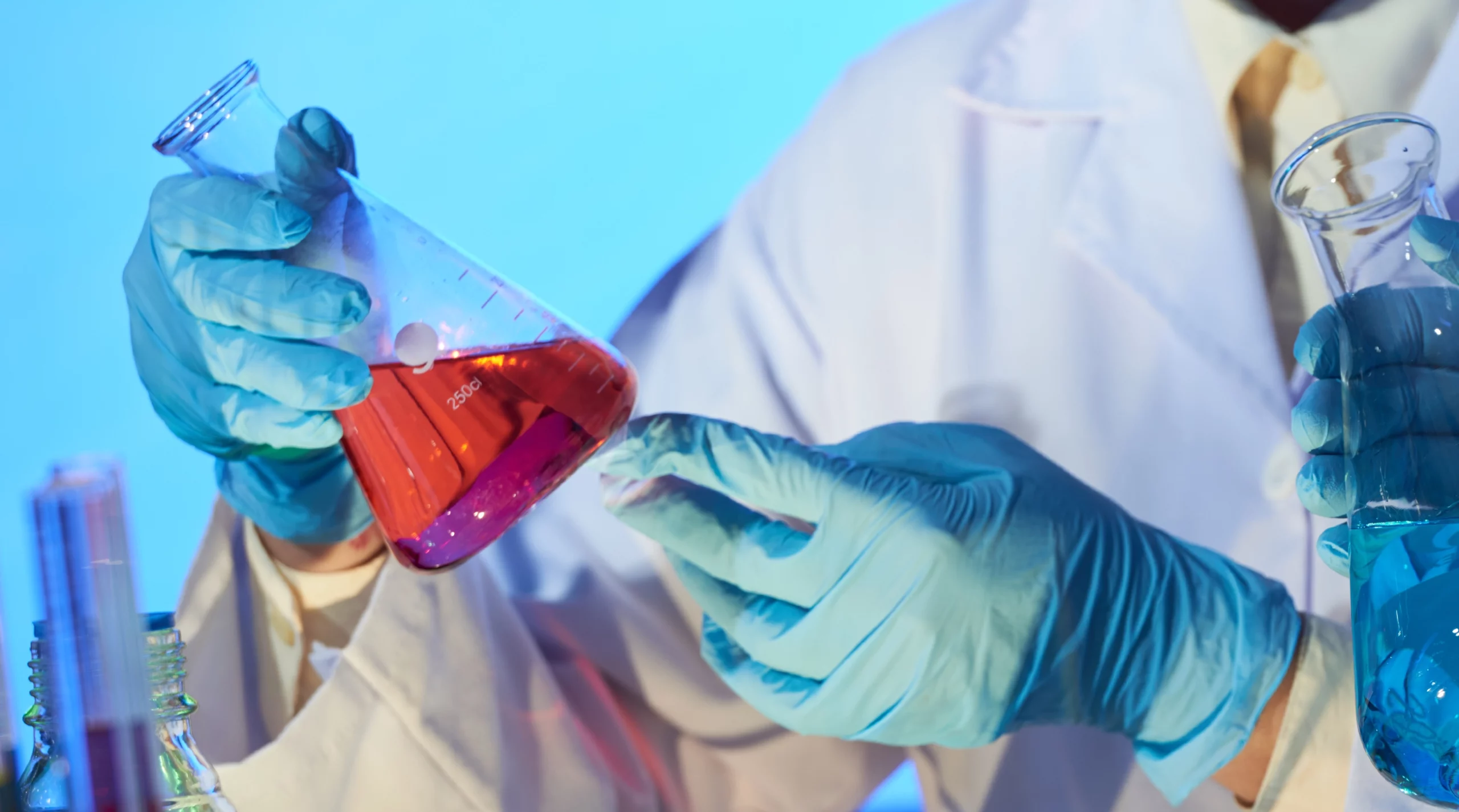 Scientifique en blouse blanche et gants bleus manipulant une fiole jaugée contenant un liquide rouge dans un laboratoire de recherche.